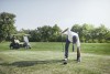 Auf dem Golfplatz, von Bäumen umrandet. im Hintergund ist ein Golfkart geparkt, im Vordergrund bückt sich ein Mann nach unten, um den Golfball aufzuheben. Er stützt sich dabei auf seinen Golfschläger.