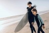 Vater und Sohn am Strand Am Strand: Ein Mann trägt unter seinem rechten Arm ein Surfboard und hält an seiner linken Hand seinen Sohn und lächelt ihn an. Im Hintergrund ist das Meer zu sehen.